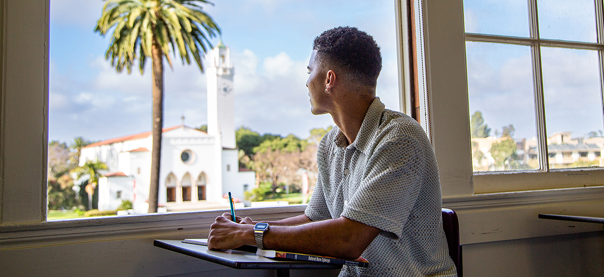 A student sitting in a classroom staring out the window with Sacred Heart Chapel and a palm tree in the background through the window.